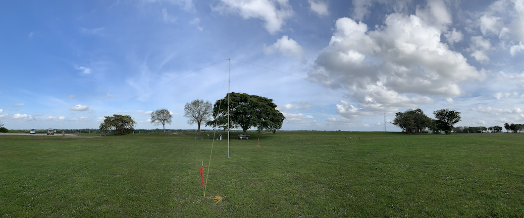 Image of an off-center-fed dipole supported by a central mast. Green grass, blue sky, clouds, and trees.
